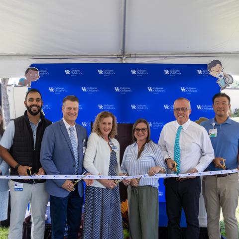 Nine people participate in a ribbon-cutting ceremony under a white tent, standing in front of a blue UK Golisano Children’s Network backdrop and holding gold ceremonial scissors as they cut a white ribbon.