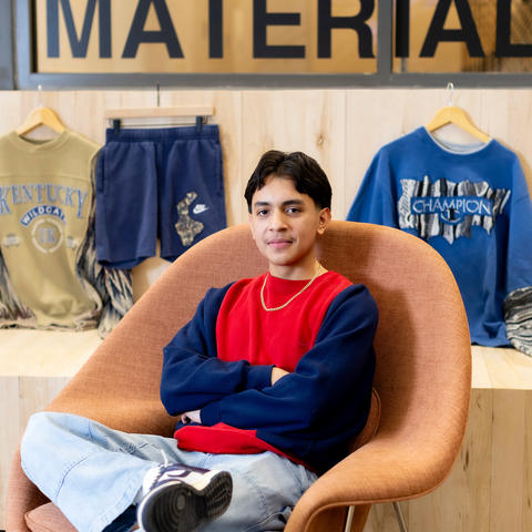 A person sits in a modern chair with arms crossed, with three graphic clothing items displayed on the wall behind them.