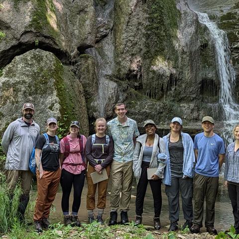 Nine people stand in front of a rocky waterfall and moss-covered cliff in a forested area.