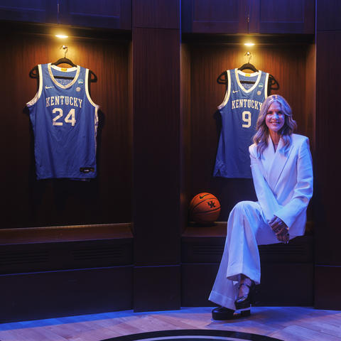 A person dressed in a light-colored suit sits in the center of a display featuring framed University of Kentucky basketball jerseys. Three blue jerseys, including numbers 24, 3 and 22, are mounted in illuminated wooden display cases behind the person.