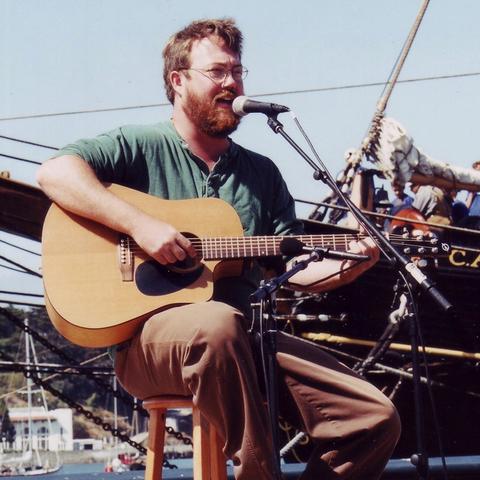 photo of Revell Carr with guitar at microphone singing sea chanteys in front of ship