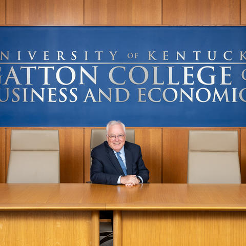 Person in a navy suit seated at a long wooden conference table beneath a blue wall sign reading “University of Kentucky Gatton College of Business and Economics,” with empty chairs on either side.