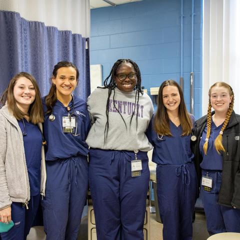 Five students stand together in a clinical setting, with exam room curtains and medical equipment visible in the background.
