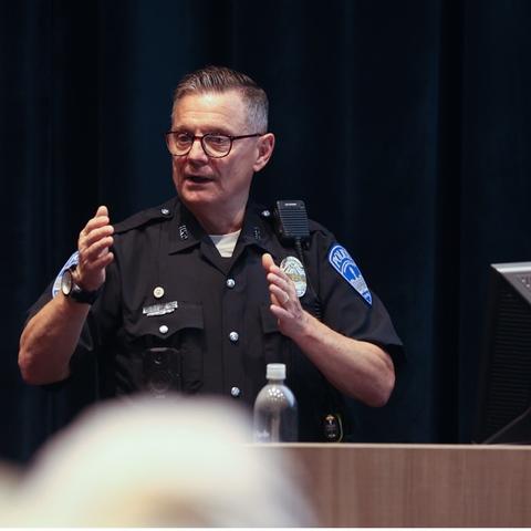 Officer Saylor teaching a staff safety course in a theater on campus. He's in uniform, talking at a lectern with his hands in the air.