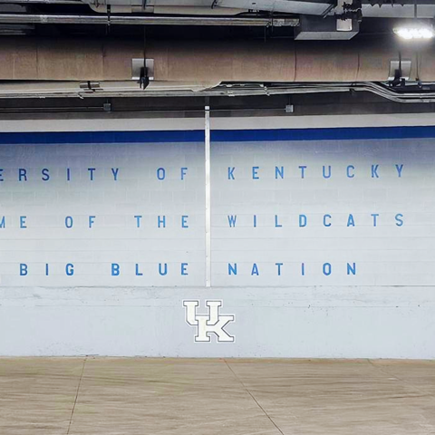 White wall inside a University of Kentucky loading dock painted with blue paw prints and the words “University of Kentucky Home of the Wildcats Big Blue Nation,” with a large UK logo centered near the floor.