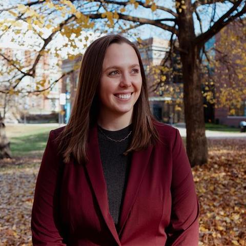 Person wearing a dark maroon blazer over a black top, standing outdoors on a campus-like setting with fallen autumn leaves on the ground.
