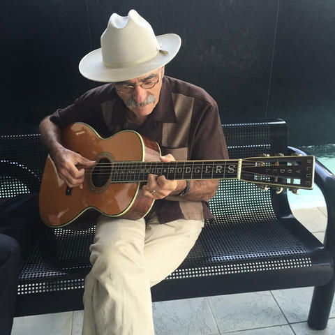 A man in a brown shirt and khaki pants sits on a bench playing guitar