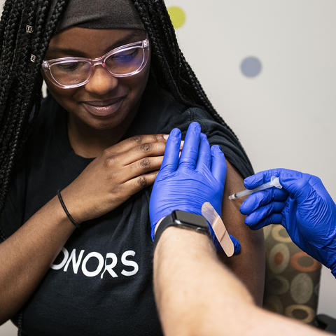 A health care worker wearing blue gloves administers a vaccine injection into a person's upper arm while the person holds up a shirt sleeve.
